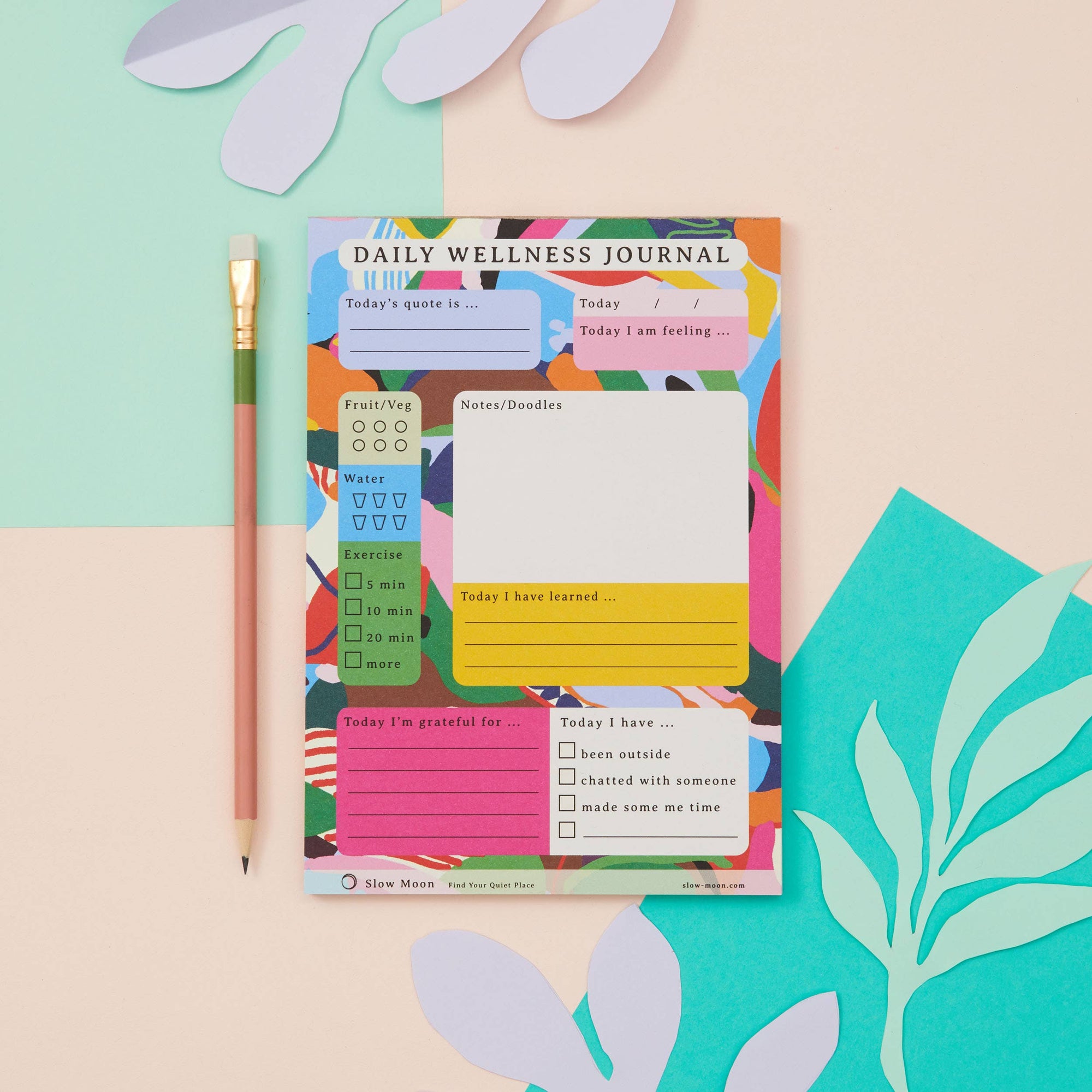 A colorful daily wellness journal with a hand-drawn Slow Moon design, placed on a flat surface accompanied by a pen and decorative elements.